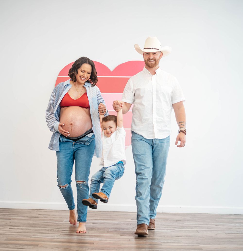 Pregnant couple swinging toddler between them in front of heart wall art indoors.