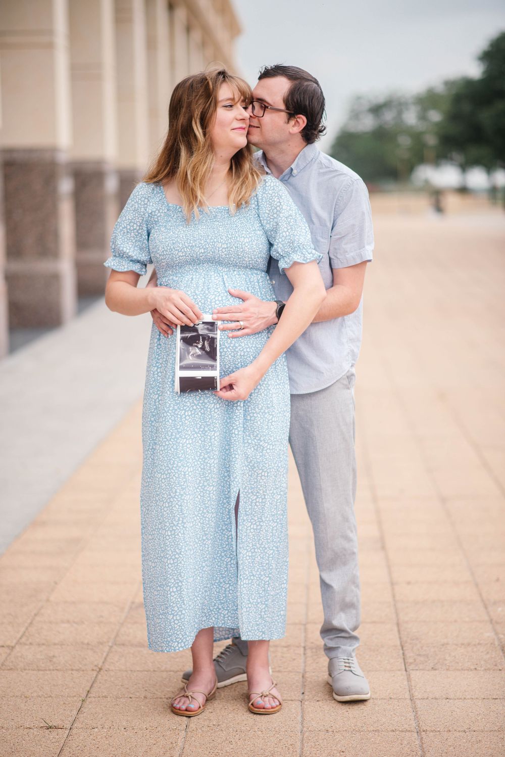 Pregnant woman in blue dress holds ultrasound photo as man kisses her cheek outdoors.