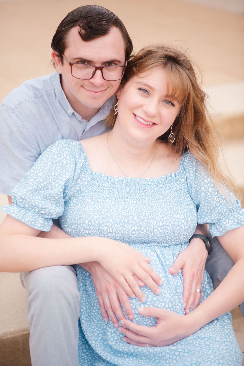 Happy couple posing for maternity photo, pregnant woman in blue floral dress with partner embracing her from behind.
