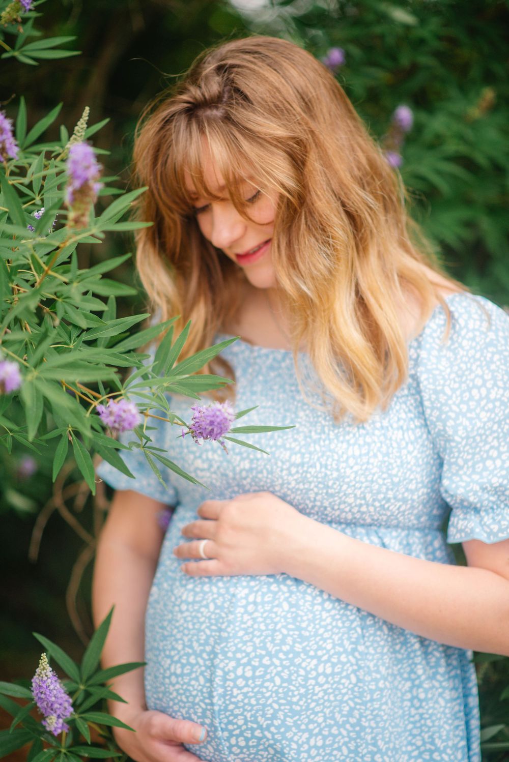 Pregnant woman in blue floral dress cradling baby bump near purple wildflowers in a lush garden setting.
