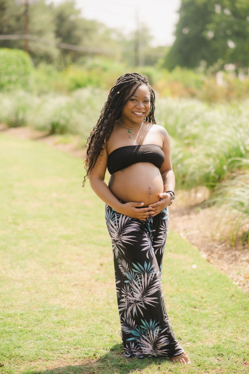 Smiling pregnant woman in black bandeau top and tropical print pants outdoors in a garden.