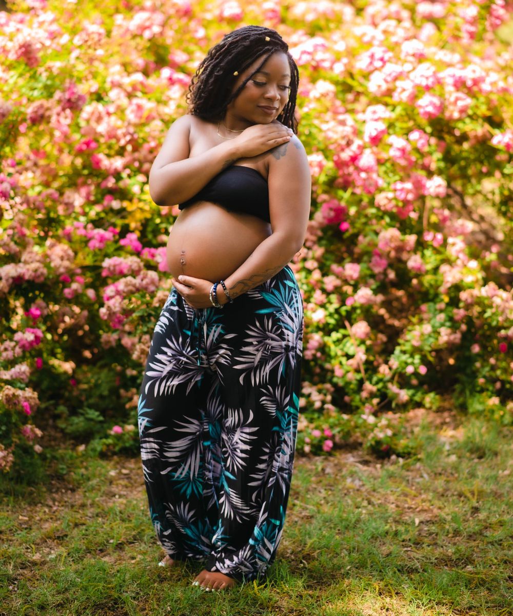 Pregnant woman in tropical print pants cradling bump in front of colorful pink flower bushes.