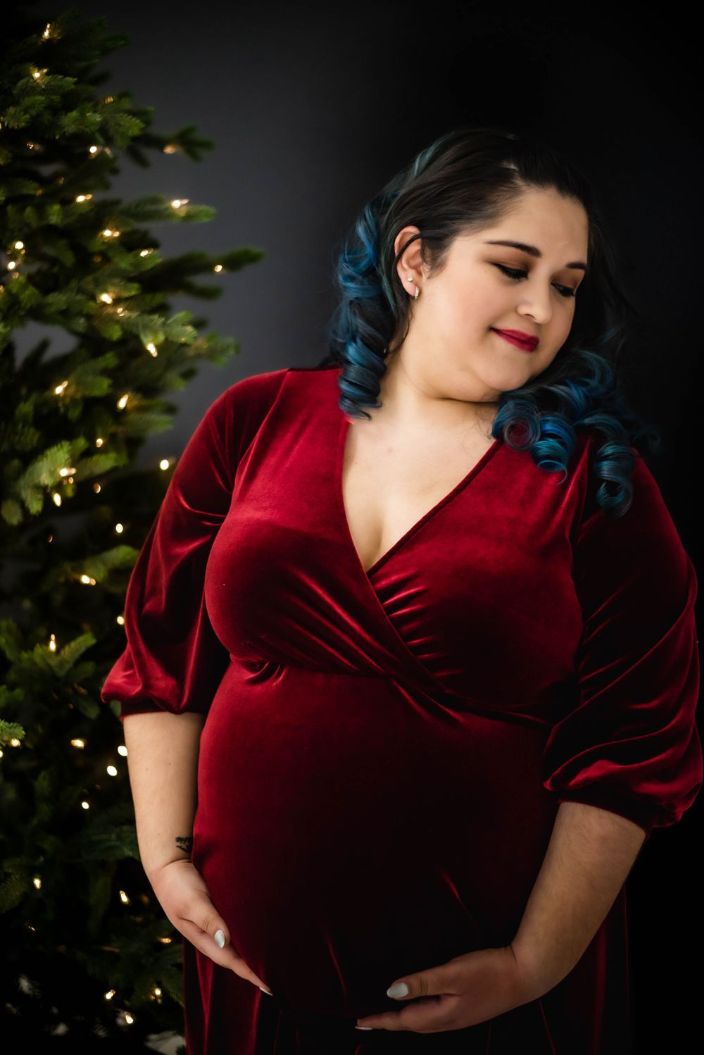 Pregnant woman in red velvet dress standing near Christmas tree with twinkling lights.