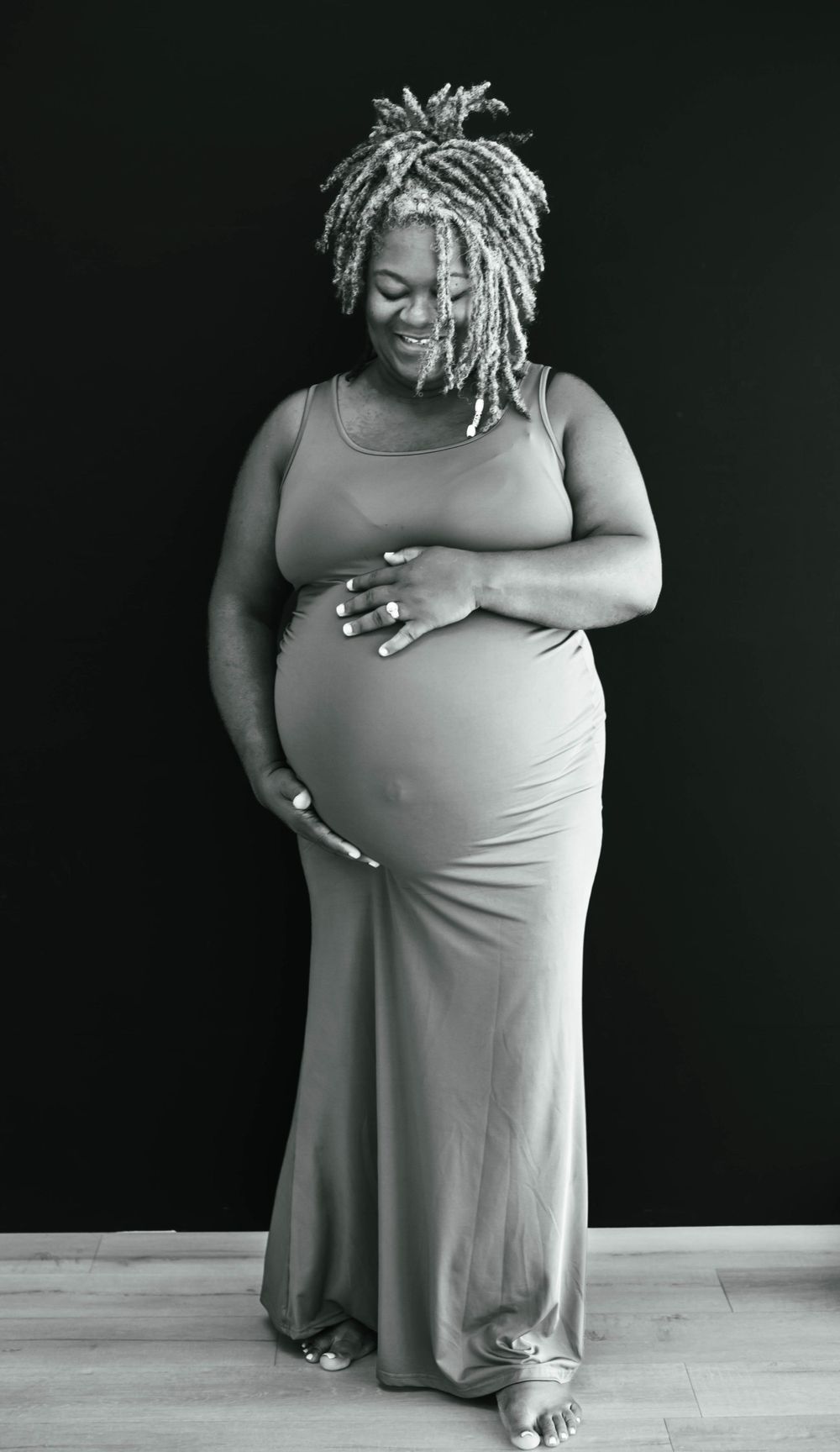 Black and white photo of smiling pregnant woman in maxi dress holding her belly.