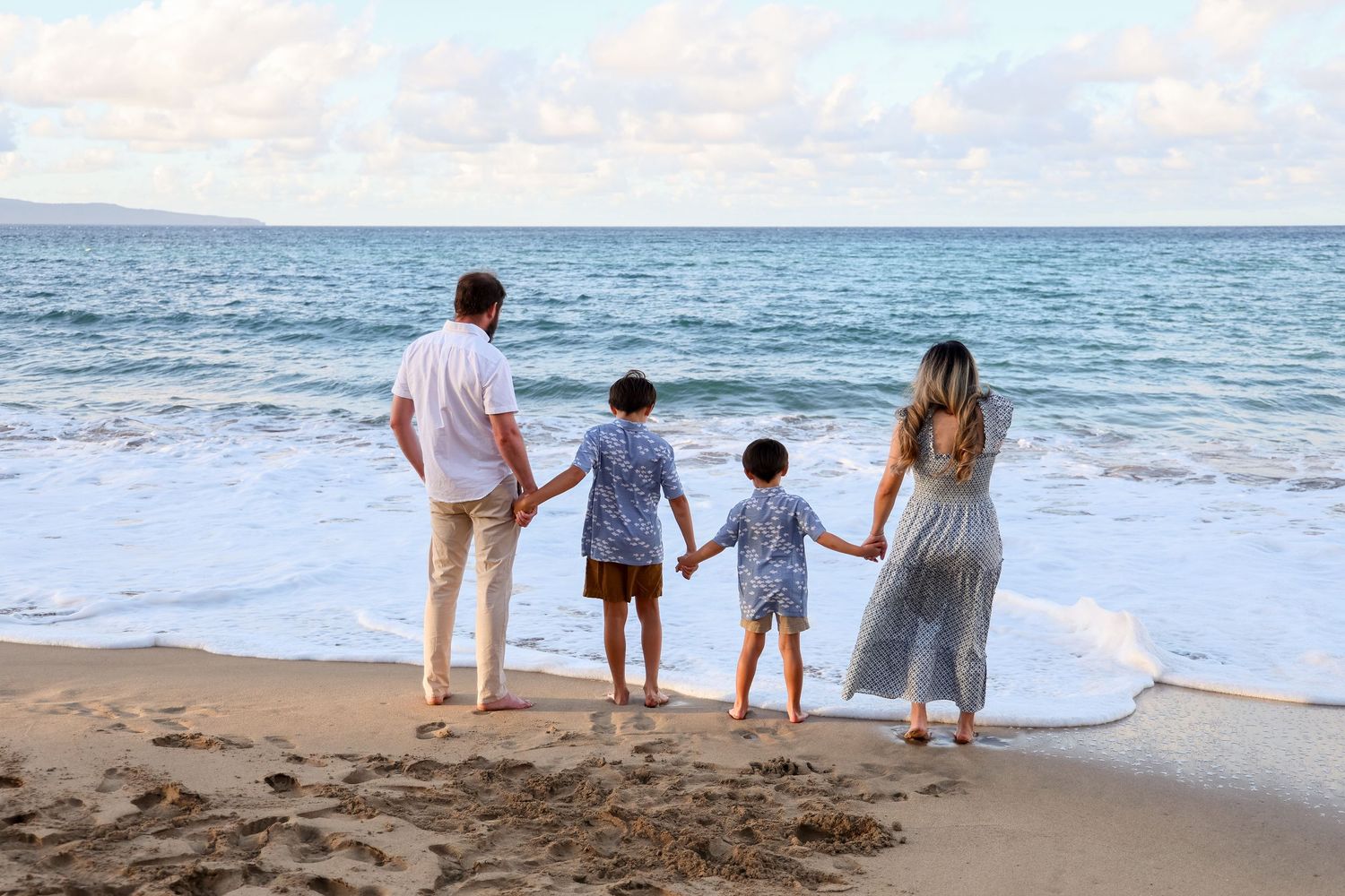 A family of four holds hands while standing at the edge of the ocean waves on a sandy beach.