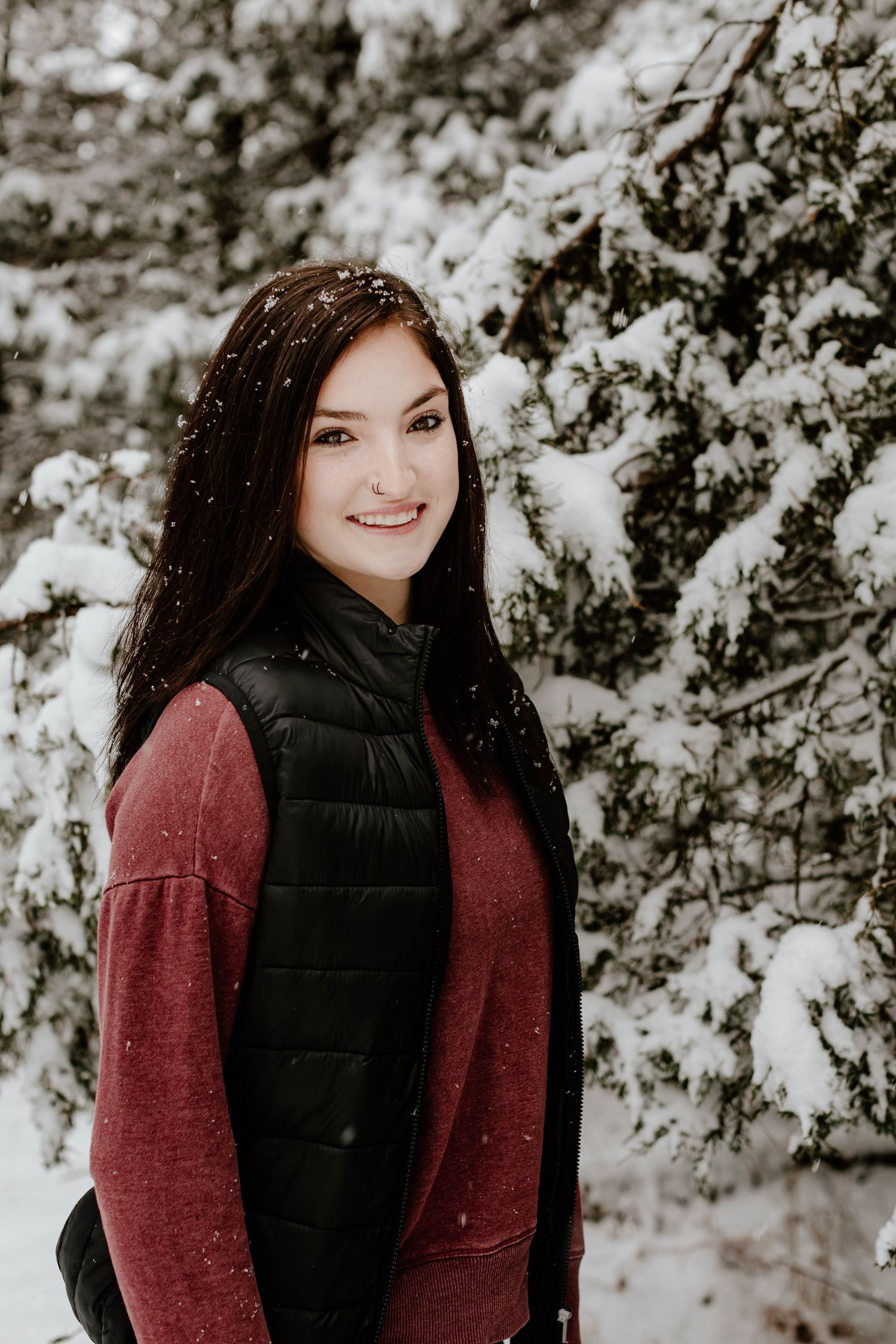 Winter portrait of person in red sweater and black vest against snowy forest background.