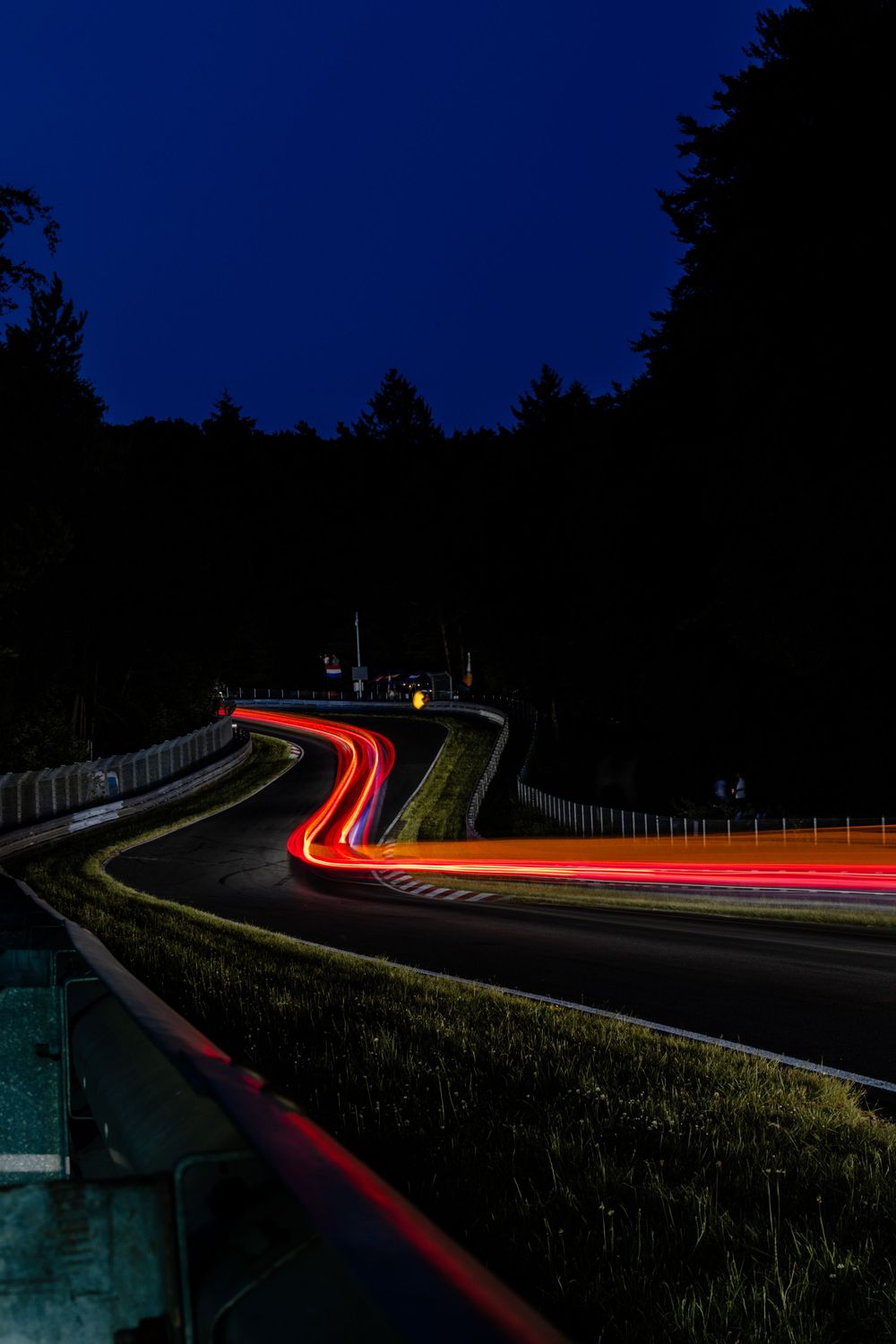 Long exposure photograph capturing red and white light trails on a dark winding mountain road at night against a deep blue sky.