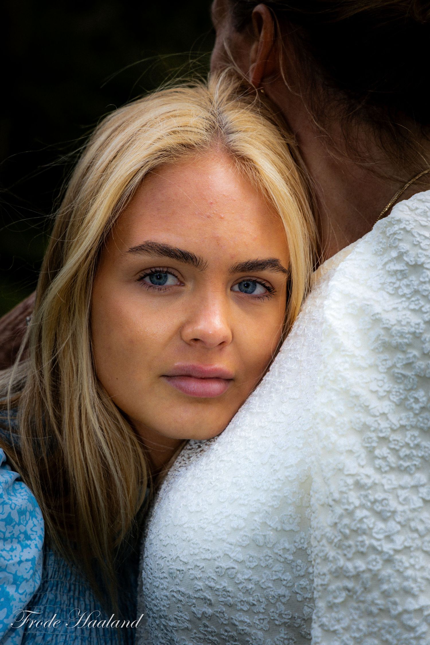 Close-up portrait of blue-eyed blonde woman resting head on someone shoulder outdoors.