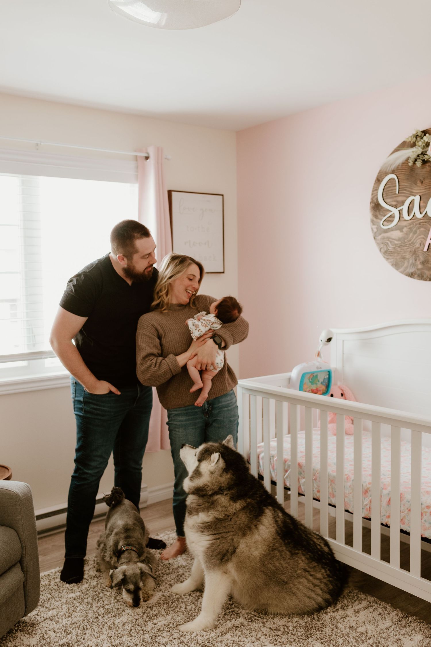 a new mom and dad holding and smiling at their baby in a pink nursery with their two dogs at their feet