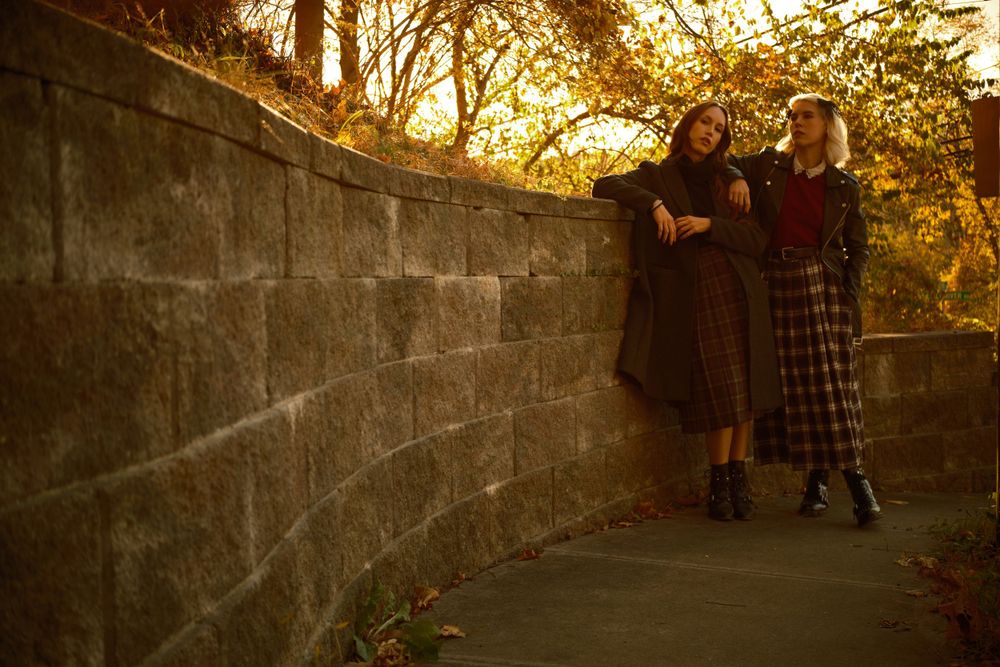 Two figures stand against stone wall during sunset in autumn setting.