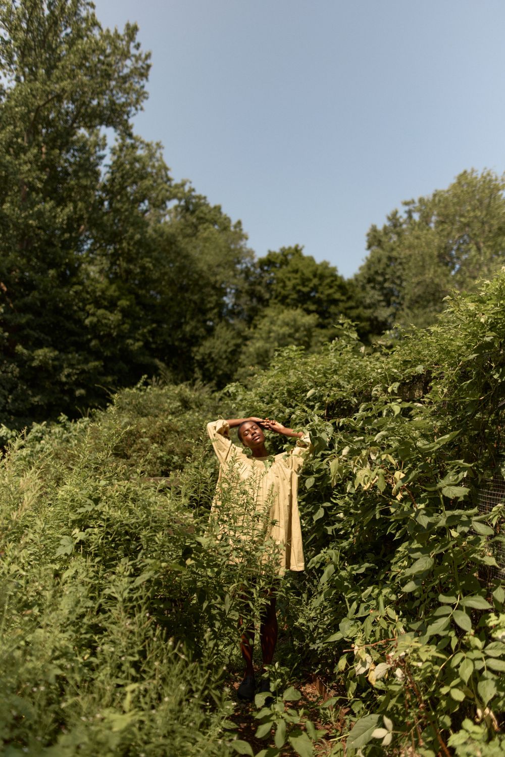 A figure in yellow clothing stands amid lush greenery against a bright blue sky.
