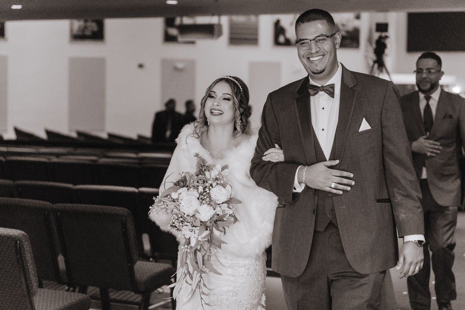 A couple walks down the aisle after their wedding ceremony in a chapel with rows of wooden pews visible.