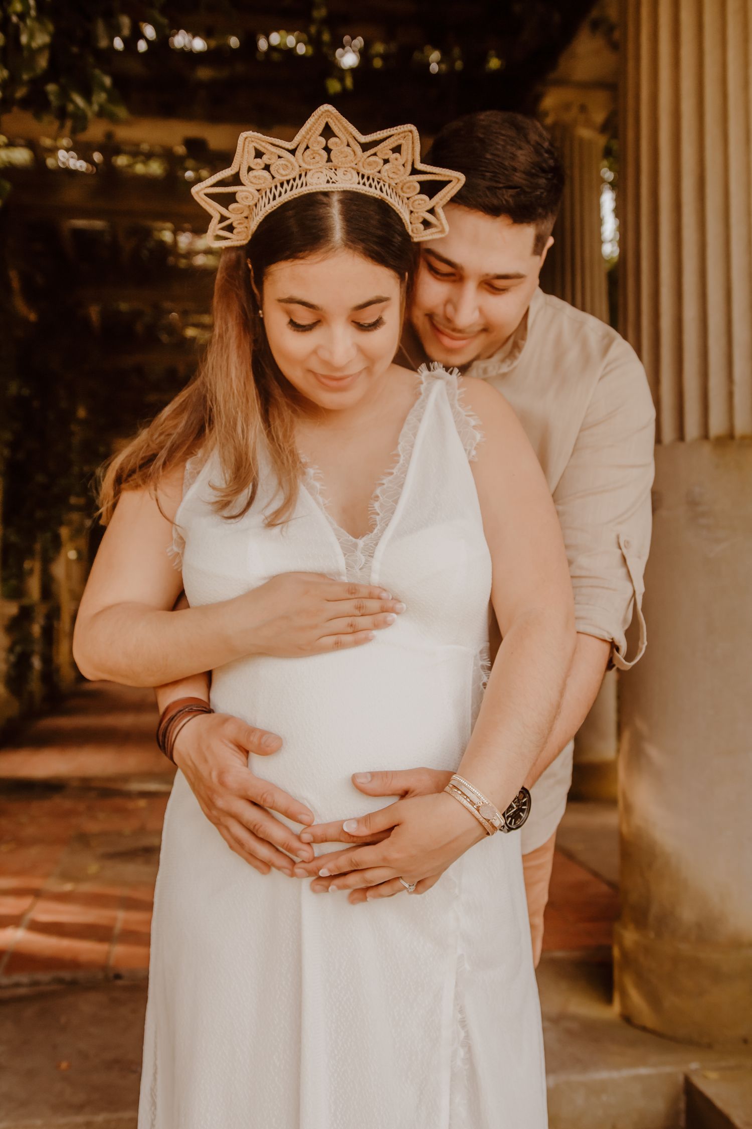 An expecting couple shares an intimate moment in warm lighting as they stand together on a porch in elegant maternity attire in Waterford.