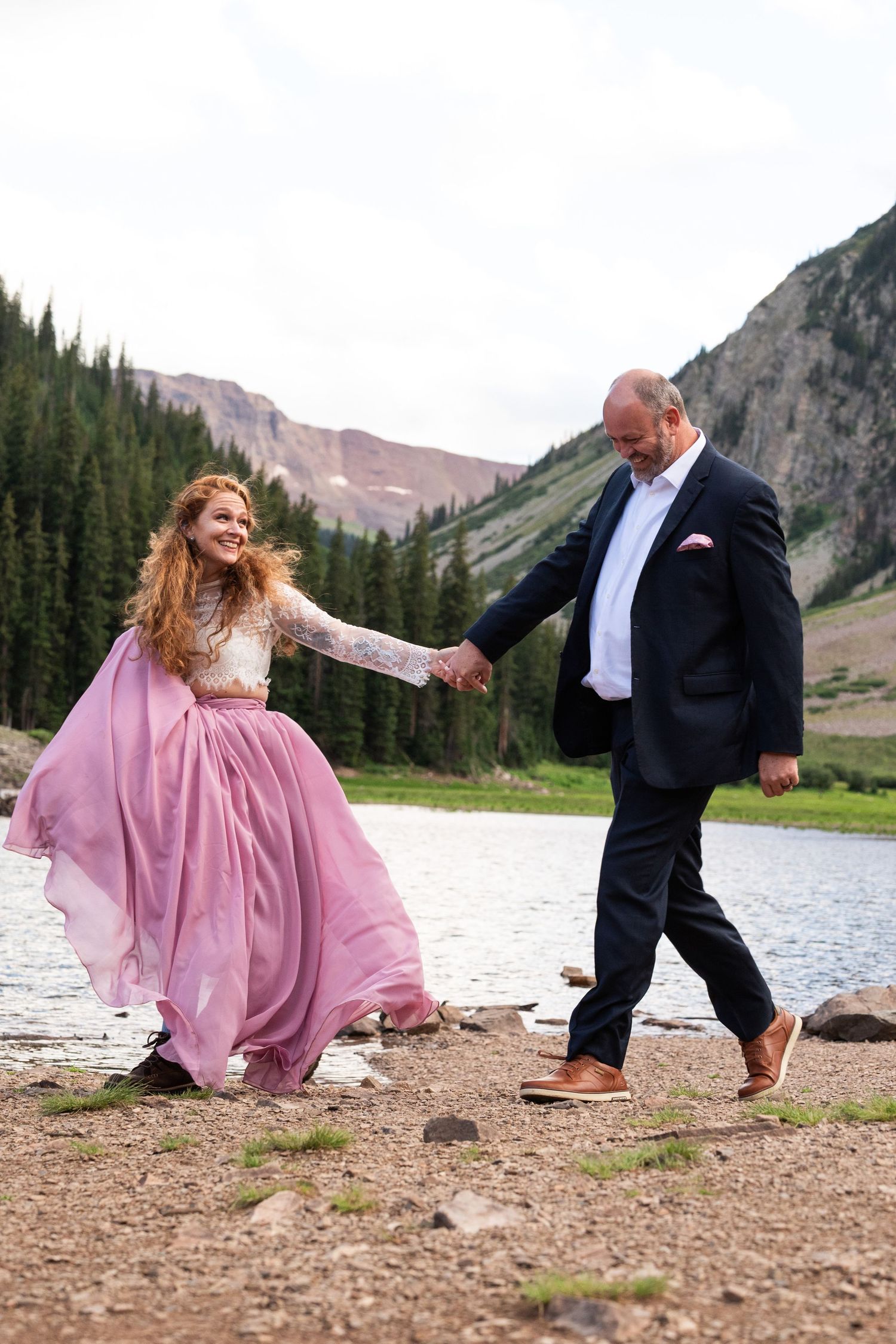 Playful couple in formal wear dancing by a mountain lake with pink flowing dress.