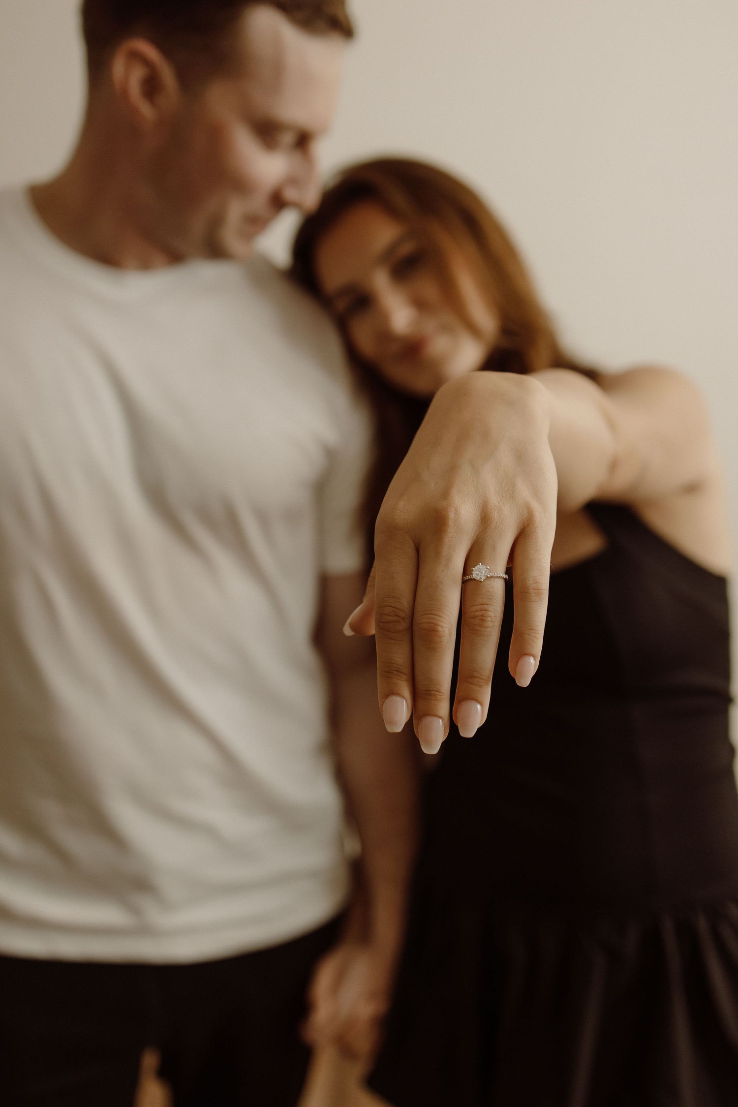 A couple in casual attire share an intimate moment while showing off a new engagement ring.