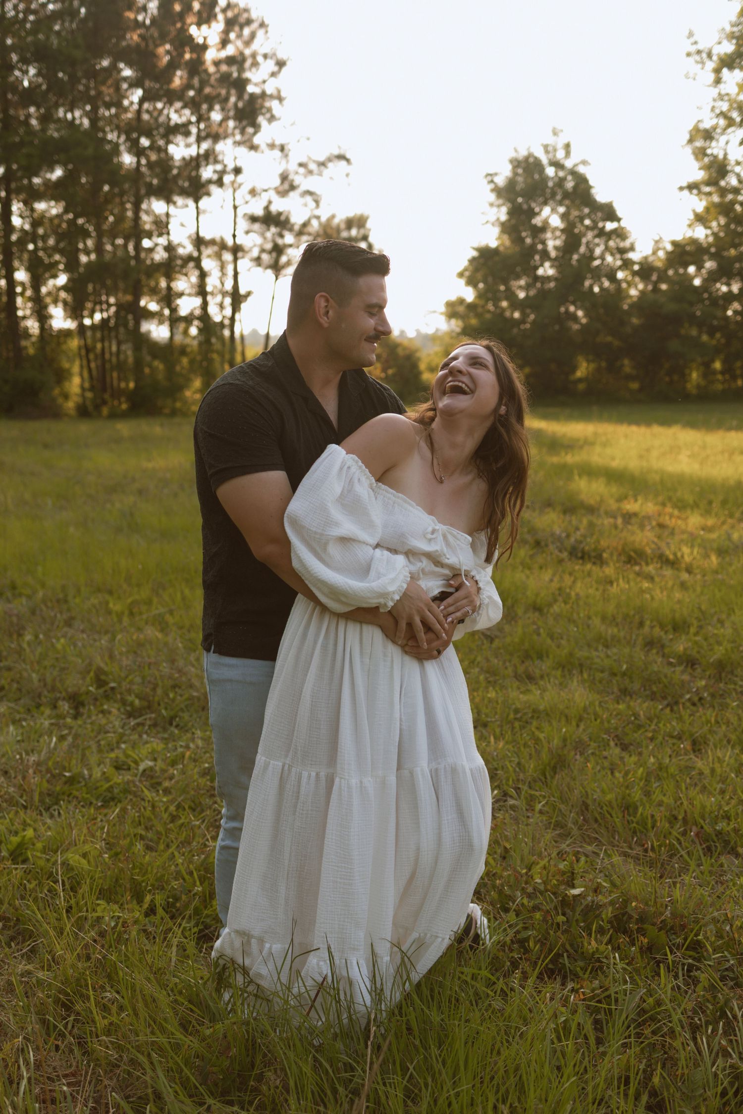A couple laughs together in a sunlit field wearing casual attire.