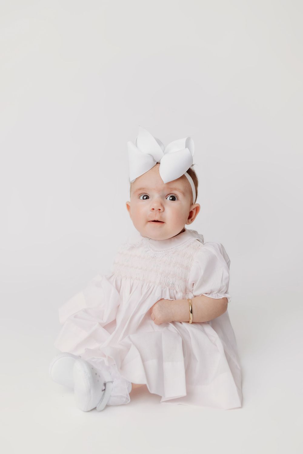 Baby in white dress and bow sitting for studio portrait against light background.