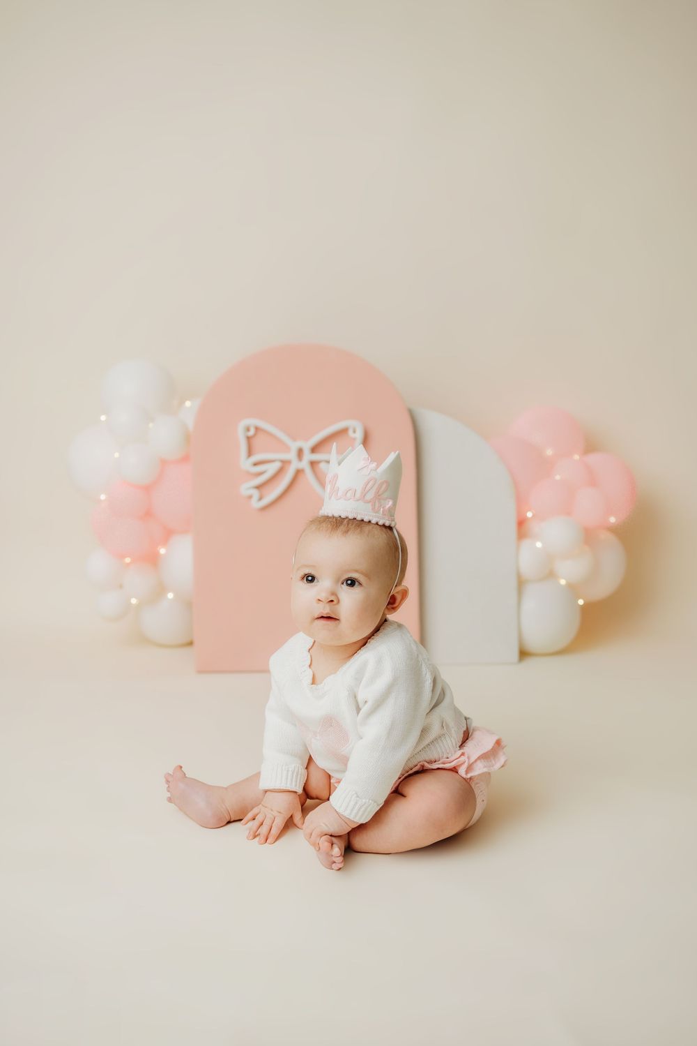 Baby in white outfit poses against pink arch backdrop with white balloon accents.