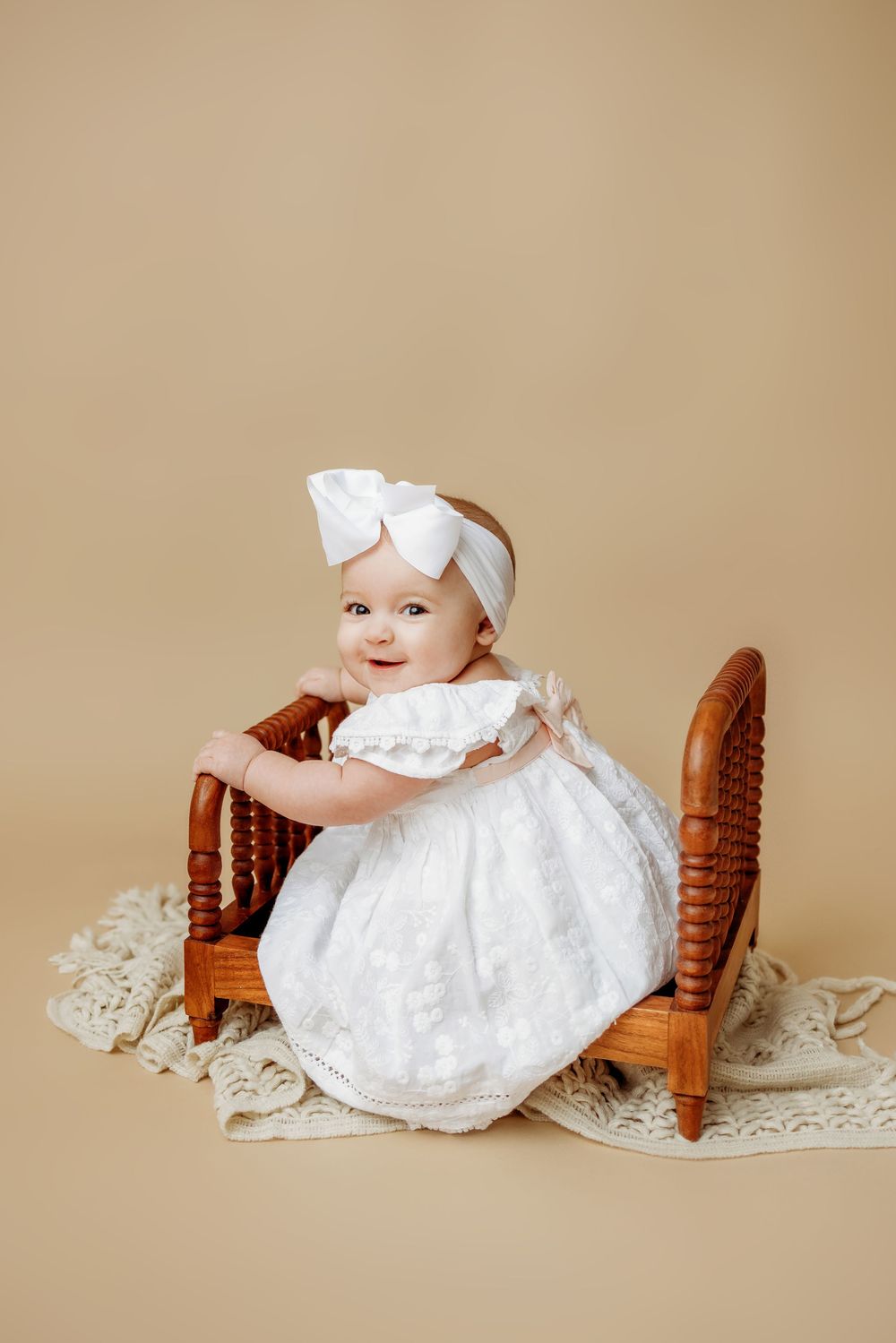A baby in white christening dress and bow sits on antique wooden furniture with lace.