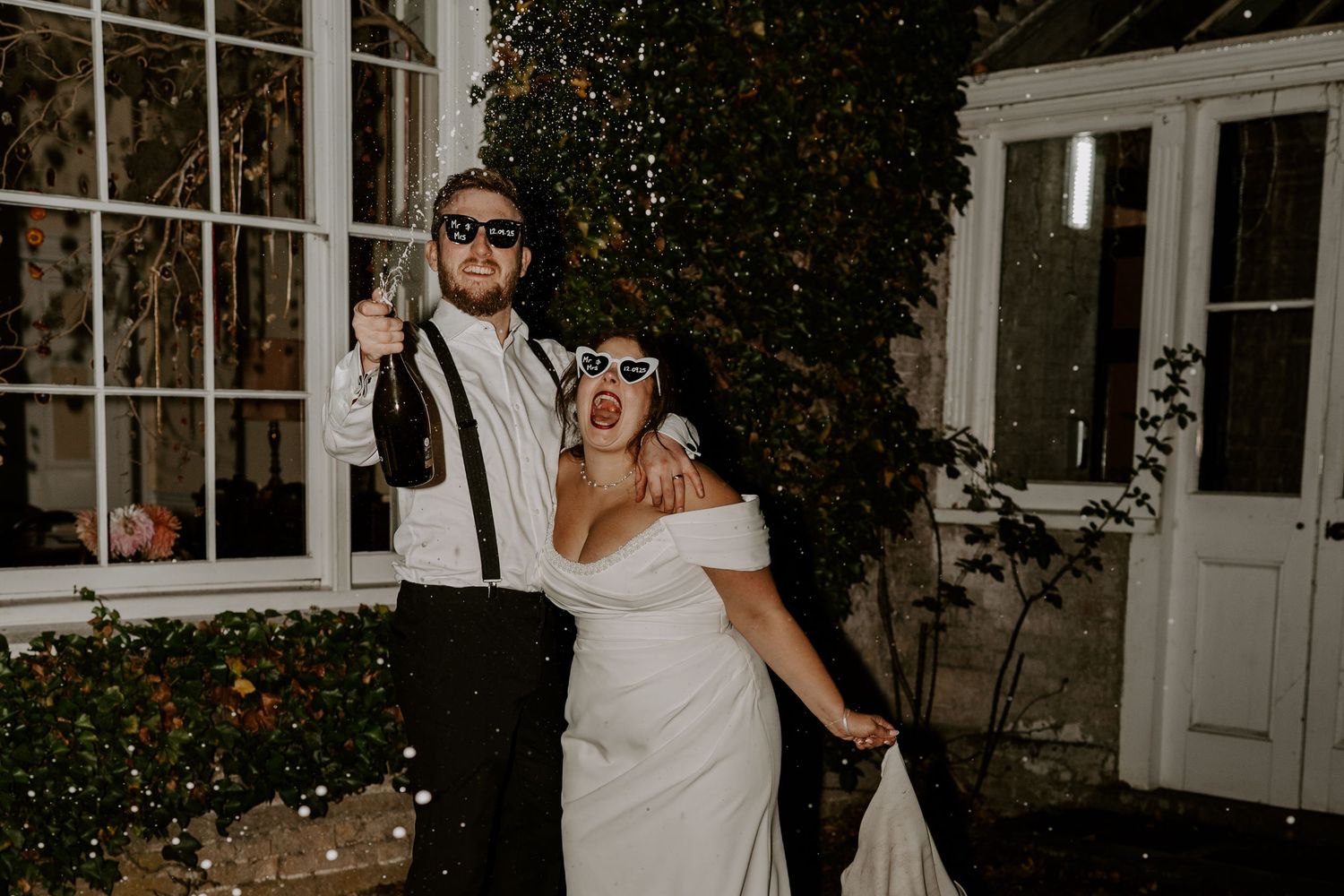 a bride and groom outside shotesham wedding venue as guests throw confetti over them. the couple are laughing and guests are cheering.