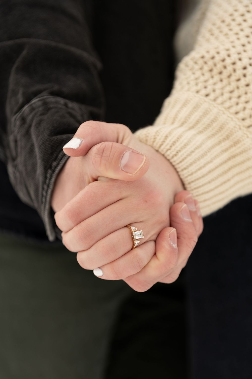 Two hands clasped together intimately, with one wearing a cream knit sweater and the other in black, showing an engagement ring.