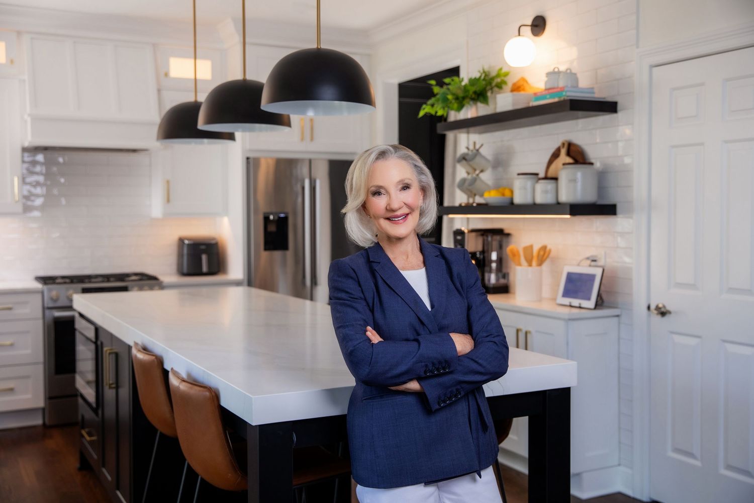 Professional stands confidently in modern white kitchen with navy blazer and crossed arms near marble island.