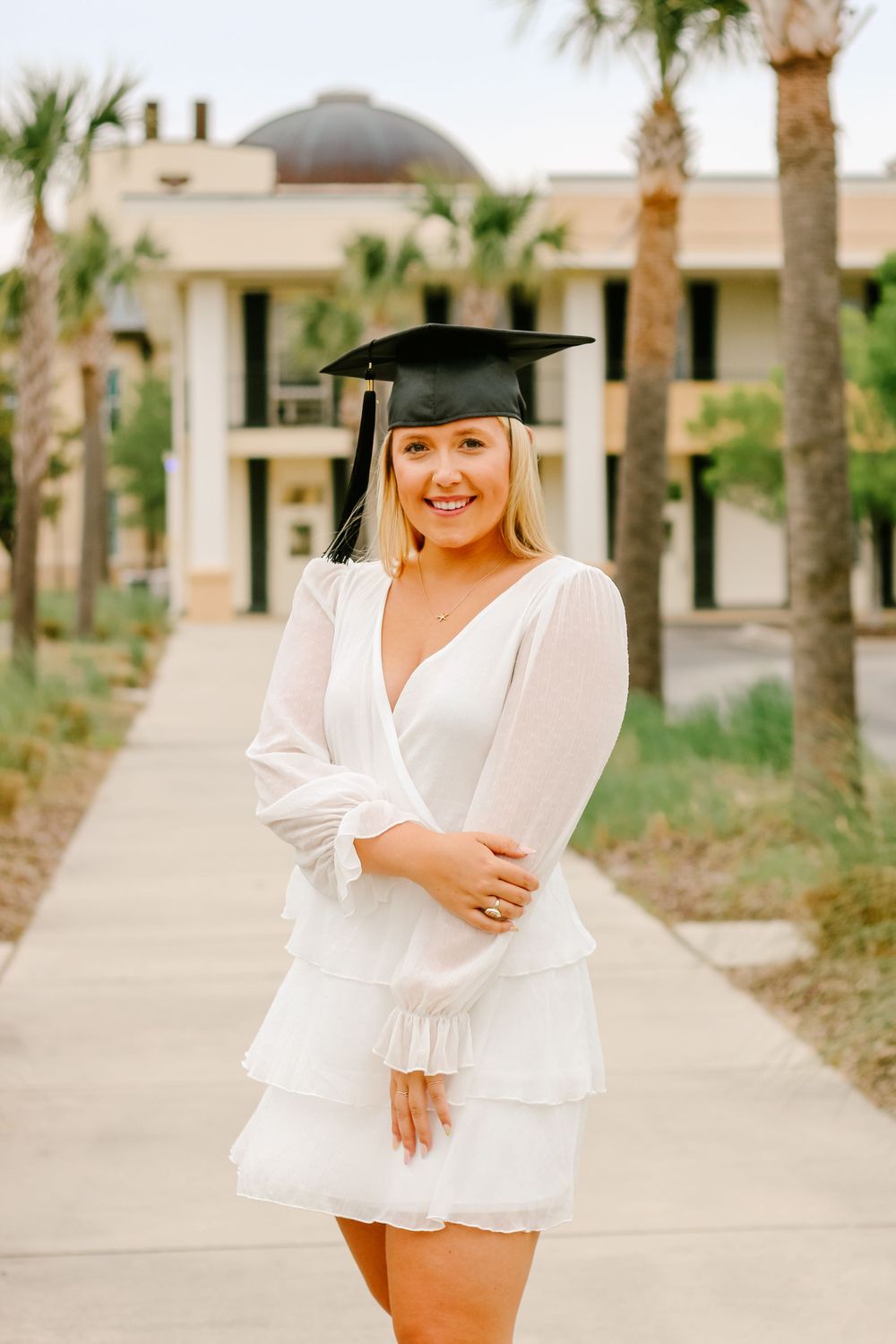 A graduate in a white dress and cap poses on a palm-tree lined campus walkway.