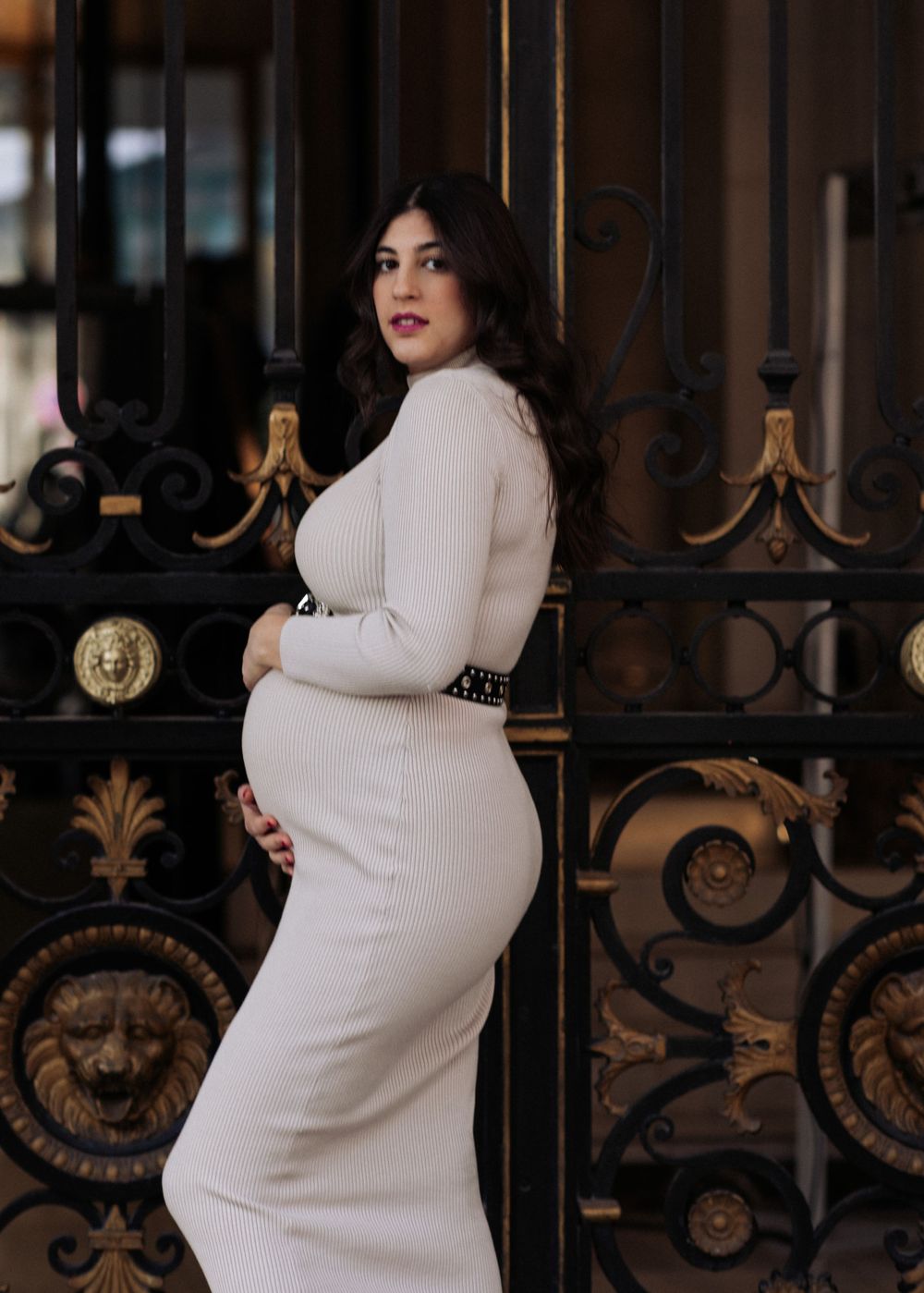 A pregnant woman in a fitted white ribbed dress poses elegantly against an ornate iron gate  in Paris
