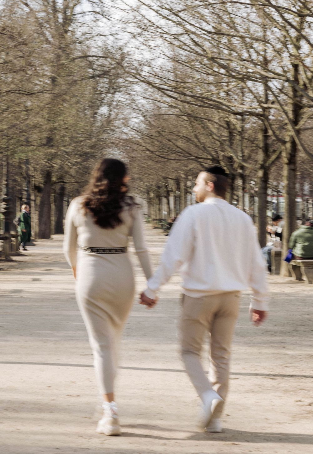 A couple holding hands while walking along a tree-lined path in matching white and cream outfits in Paris