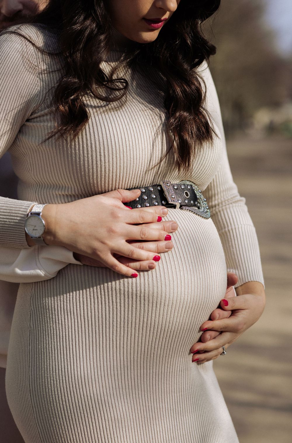 Close-up view of pregnant belly in cream ribbed sweater with hands showing red nail polish resting on the bump.