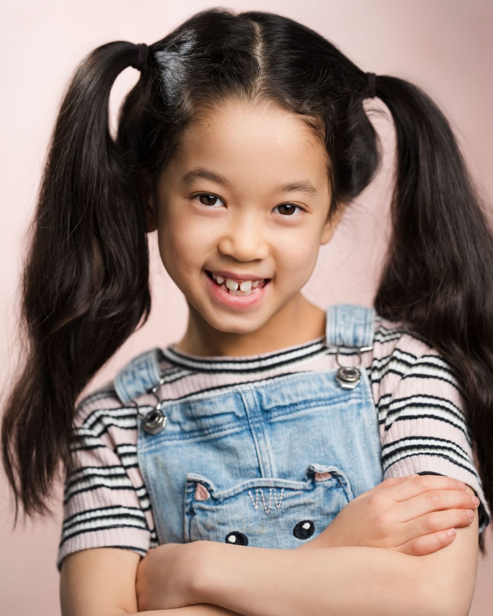 Young child wearing denim overalls and striped shirt with pigtails smiling at camera against pink background. Children's headshots Surrey.