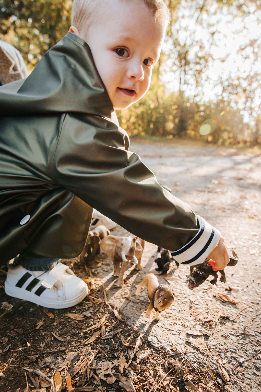 Small hands in white sneakers explore fallen autumn leaves and mulch on the ground in warm sunlight.