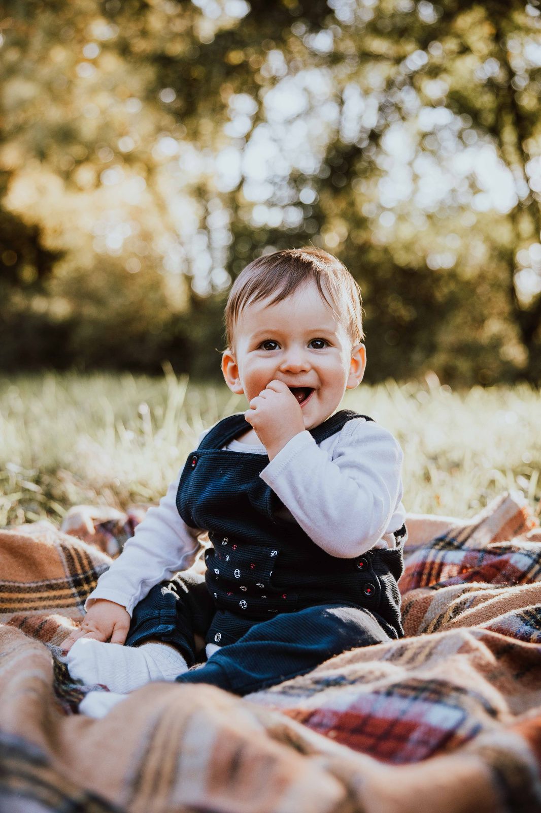 Baby in navy blue overalls lies on plaid blanket outdoors while smiling and playing in autumn sunlight.