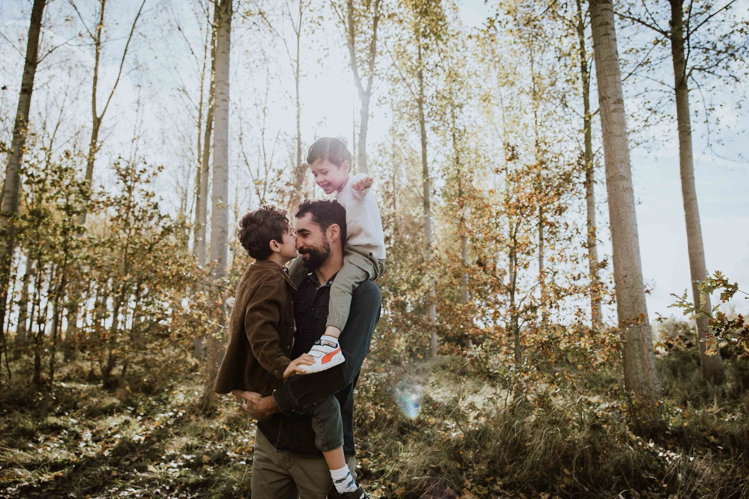 A couple embraces in a sunlit forest during autumn with tall trees and golden light filtering through the branches.