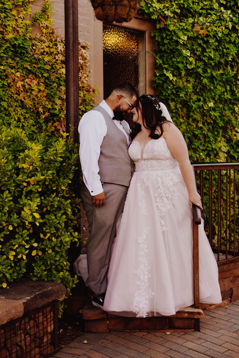 A romantic couple in wedding attire share a tender moment on brick steps surrounded by lush green ivy.