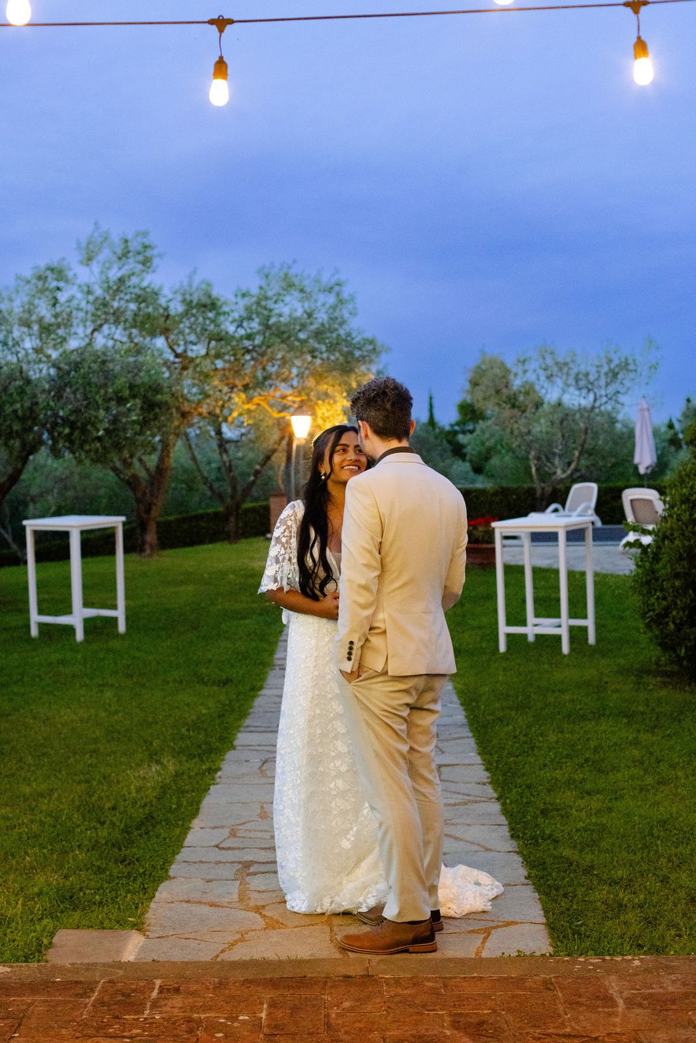 A couple in wedding attire shares an intimate moment on a stone path under string lights at dusk in an outdoor garden setting.