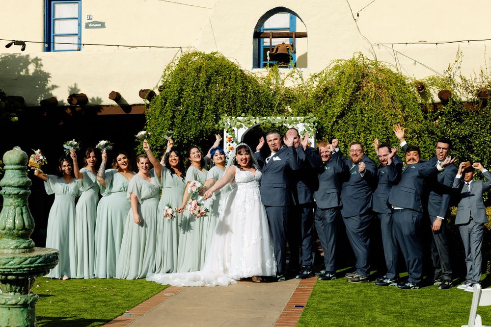 Wedding party in formal attire stands together on a sunny lawn during an outdoor celebration.