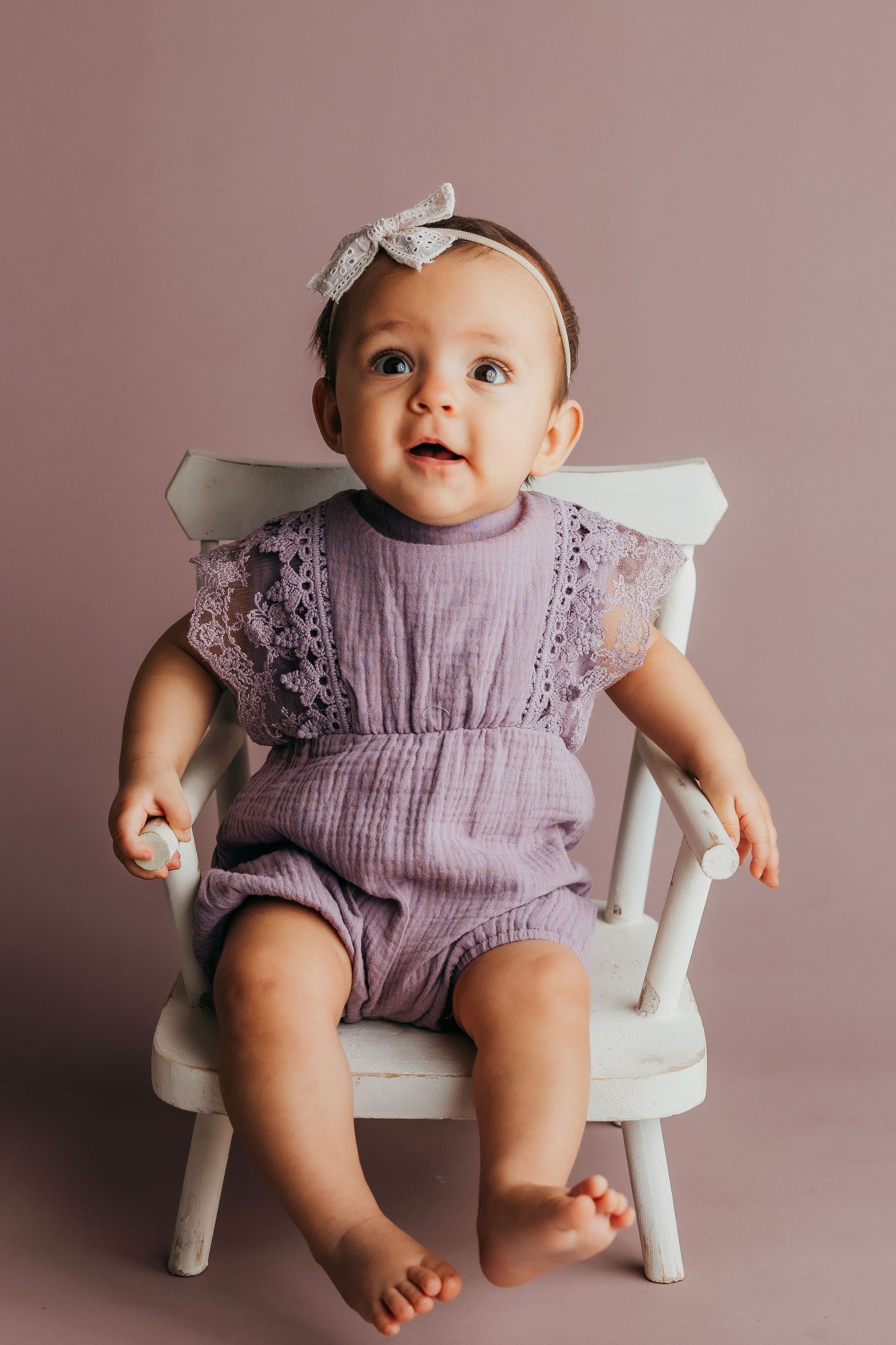 Baby in peach dress and bow headband sitting on white floor.