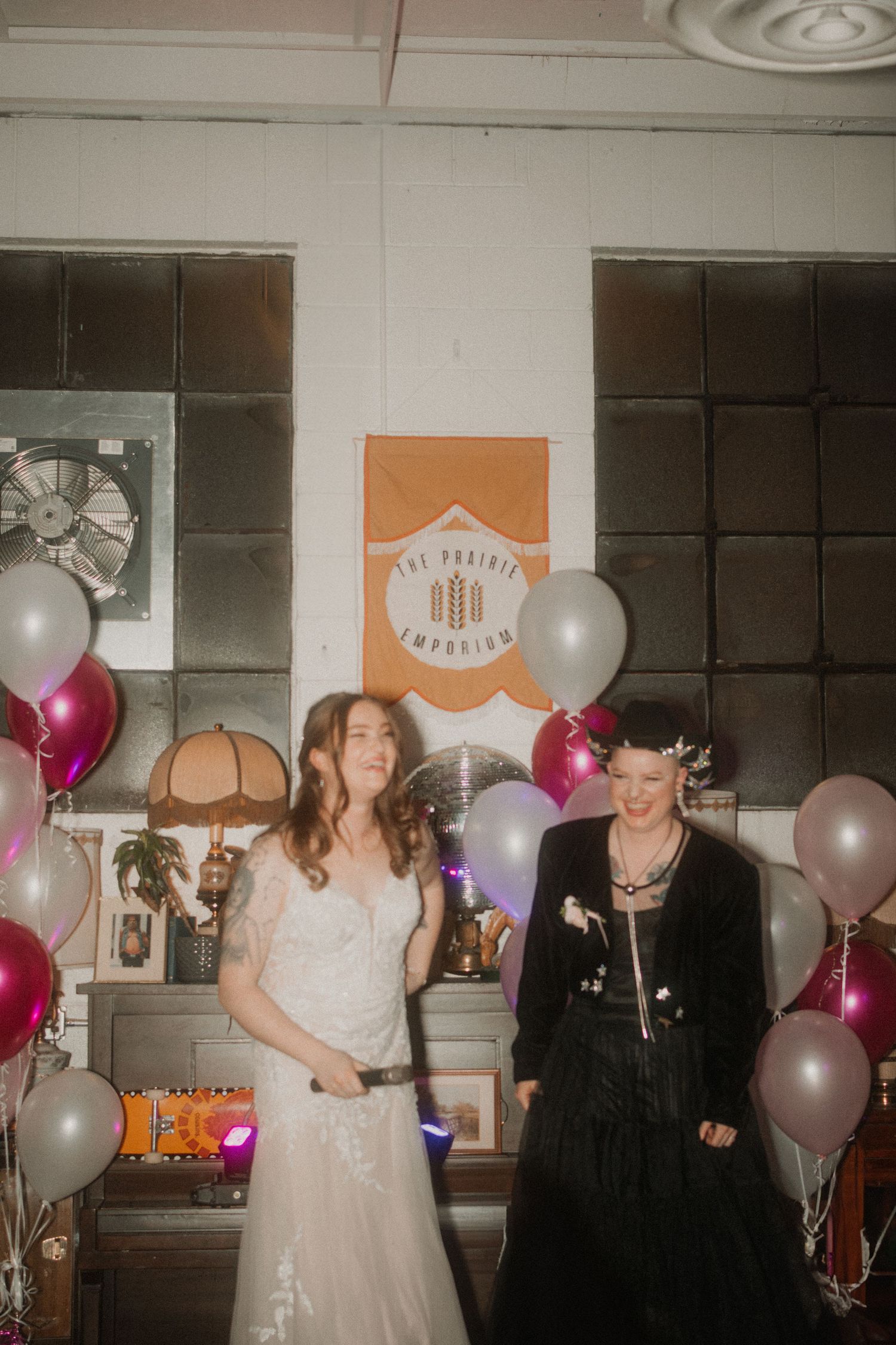 Two people in contrasting white and black outfits pose at a celebration with balloons and decorations.