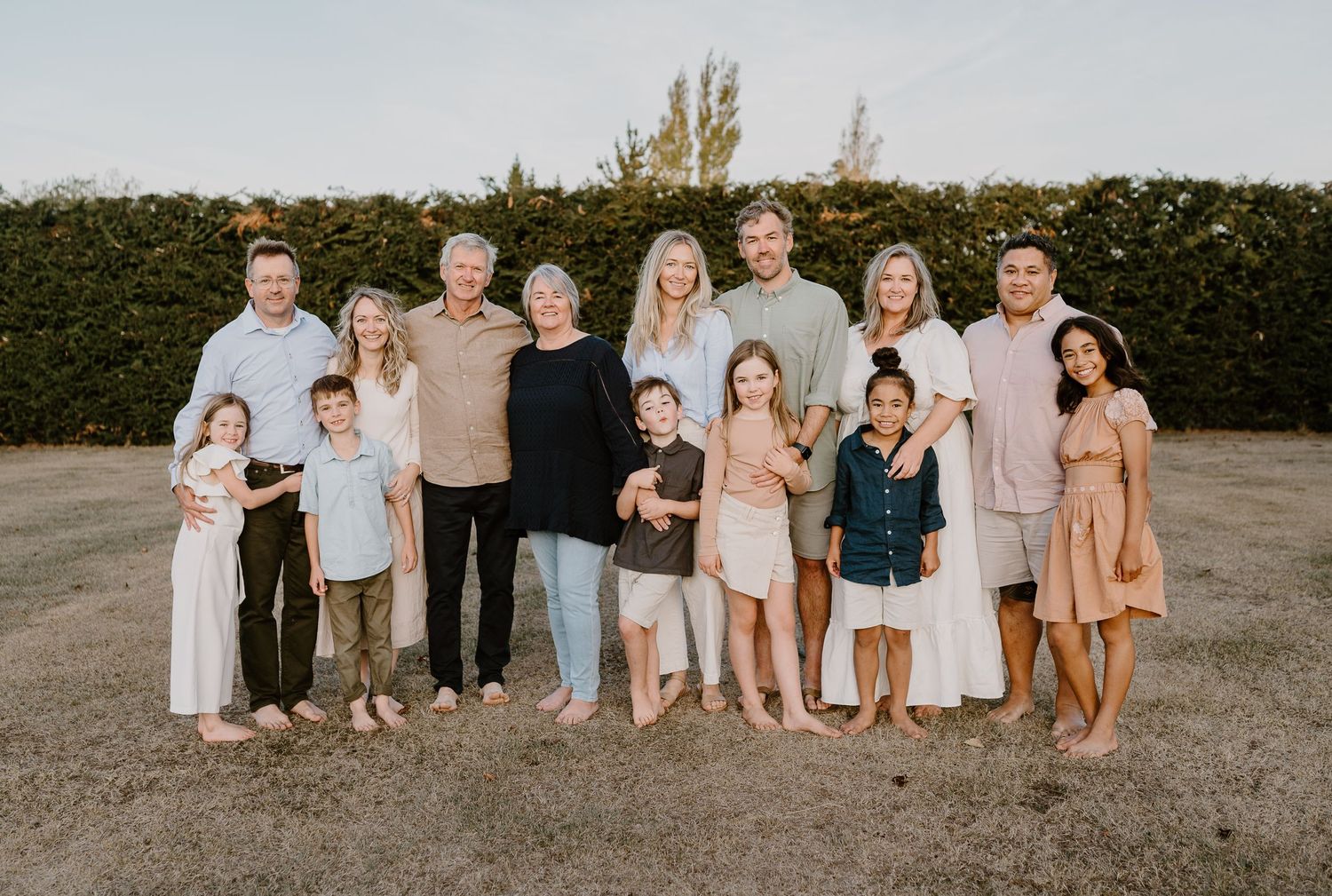 Extended family group poses together outdoors against a tall hedge during golden hour.