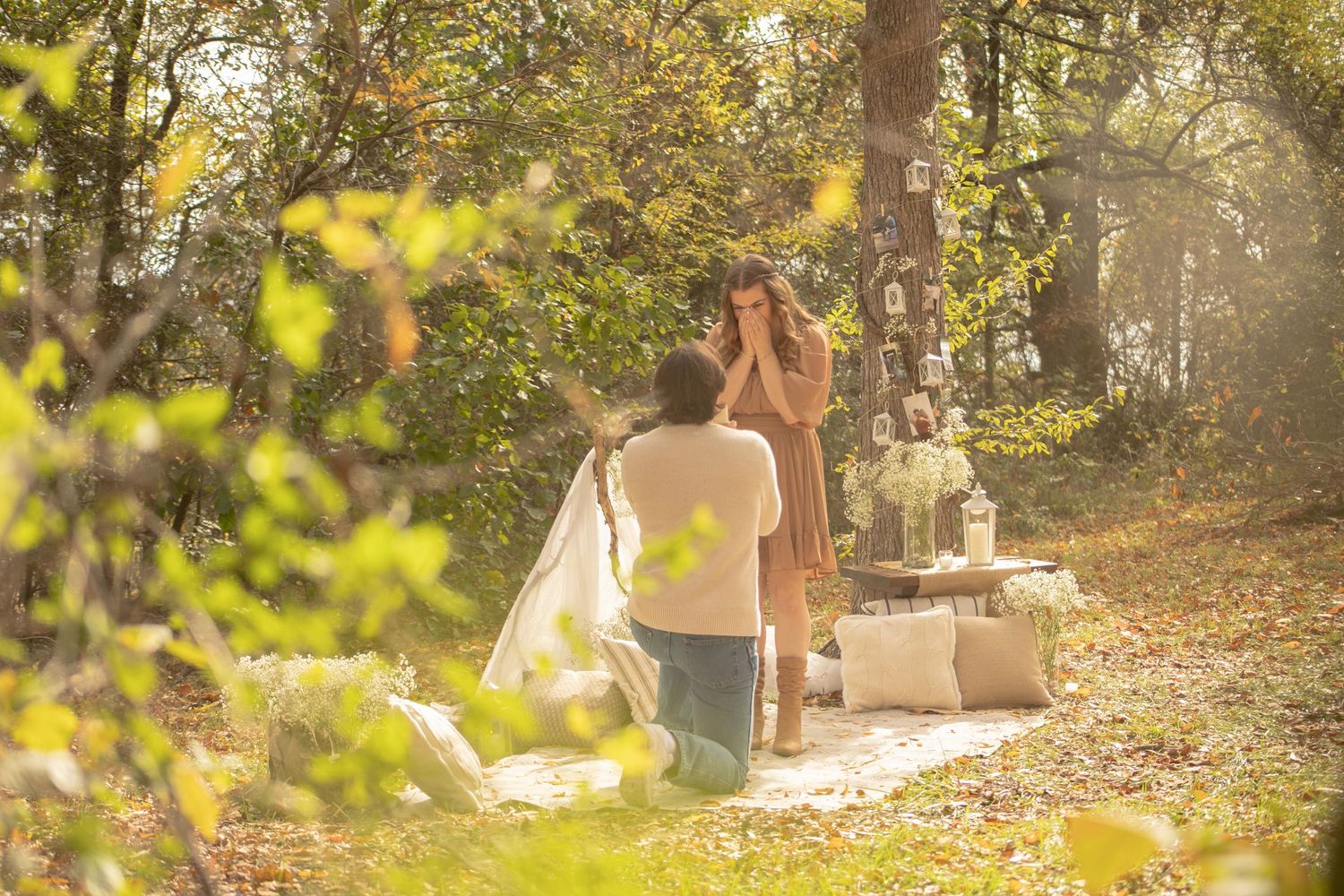 Man kneeling to propose to woman in a decorated woodland setting with lanterns and flowers.