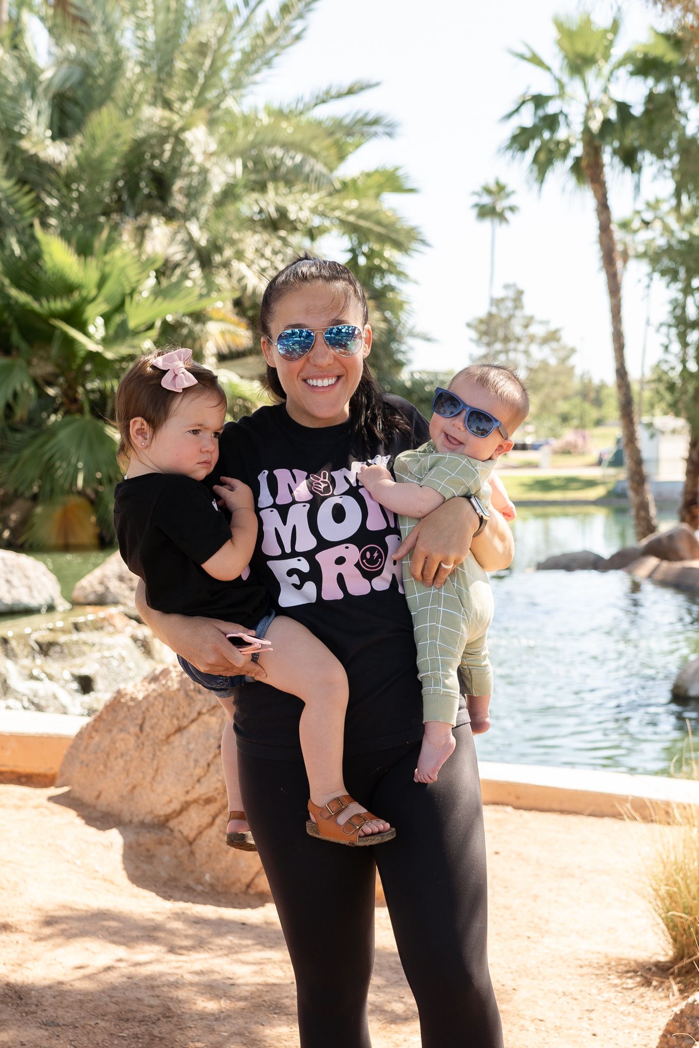 A person in sunglasses holds two small children by a waterfront with palm trees in the background.