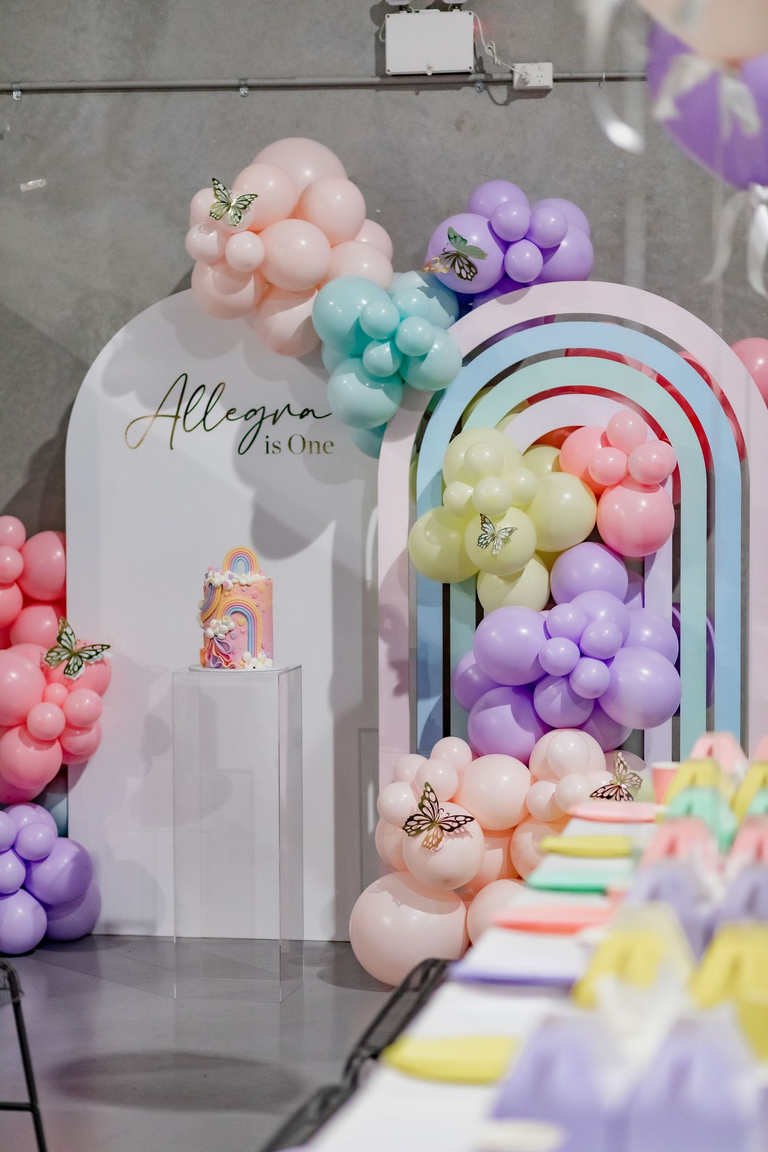 Pastel balloon arch installation with butterflies and 'Allegra is One' signage in an industrial space.