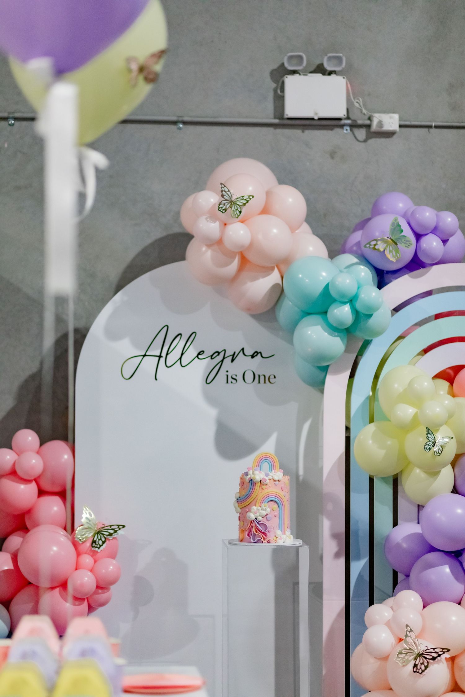 Elegant balloon arch display with 'Allegra is One' text and pastel colored balloon decorations.