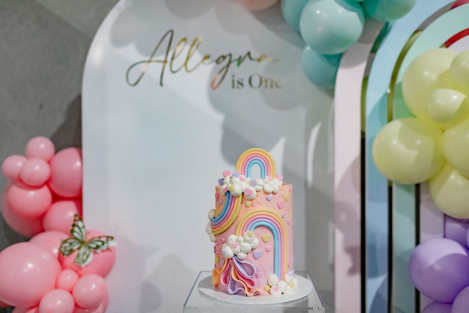 Close-up of a whimsical rainbow birthday cake under a frosted dome with 'Allegra is One' text.