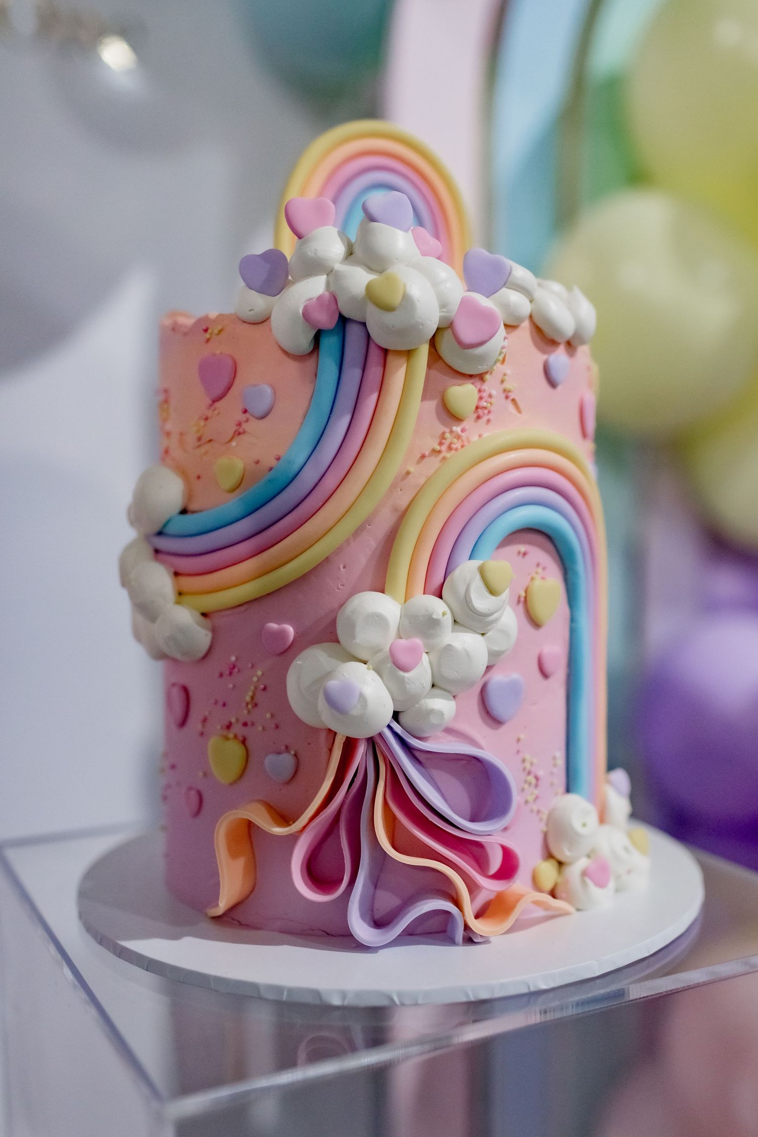 A pink fondant cake decorated with colorful rainbow designs, clouds, and pastel accents.