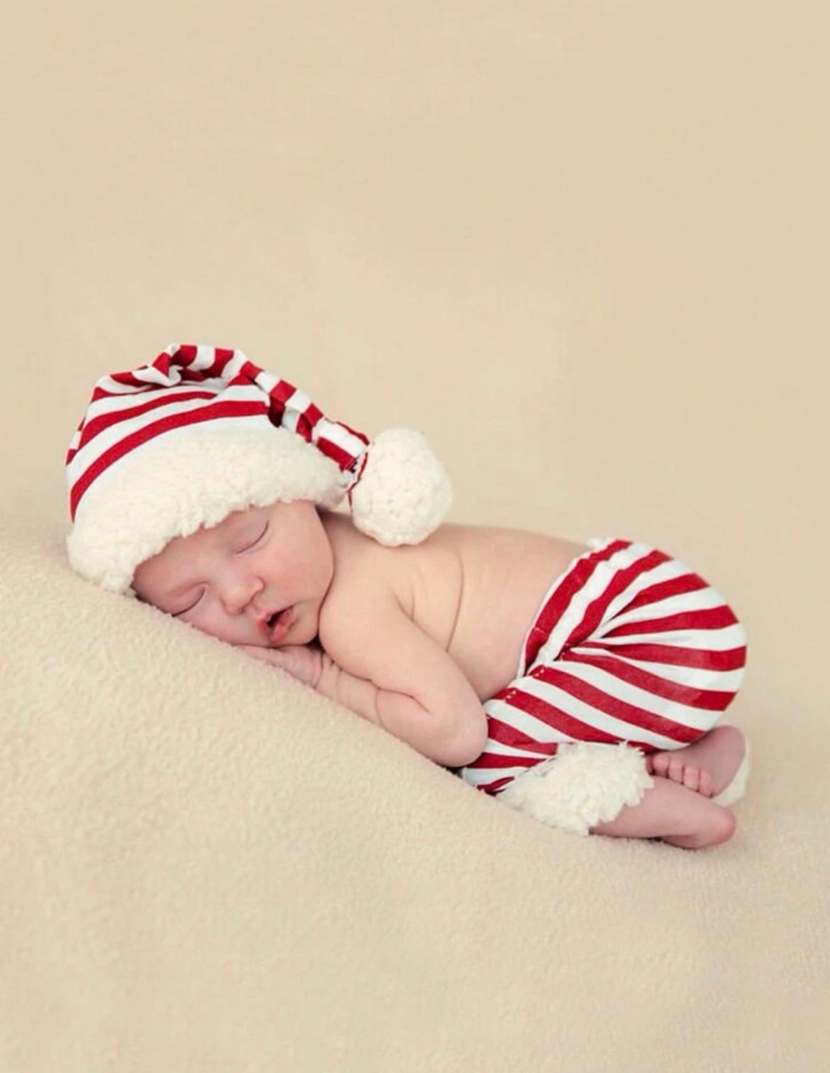 Sleeping newborn wearing red and white striped holiday outfit on cream background.