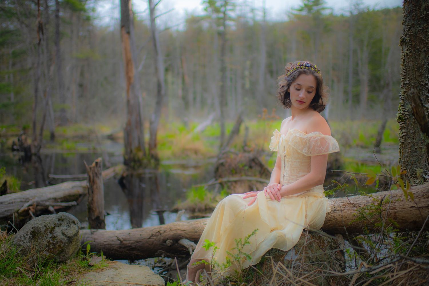 Woman in vintage cream dress sits dreamily on fallen log in misty forest swamp.