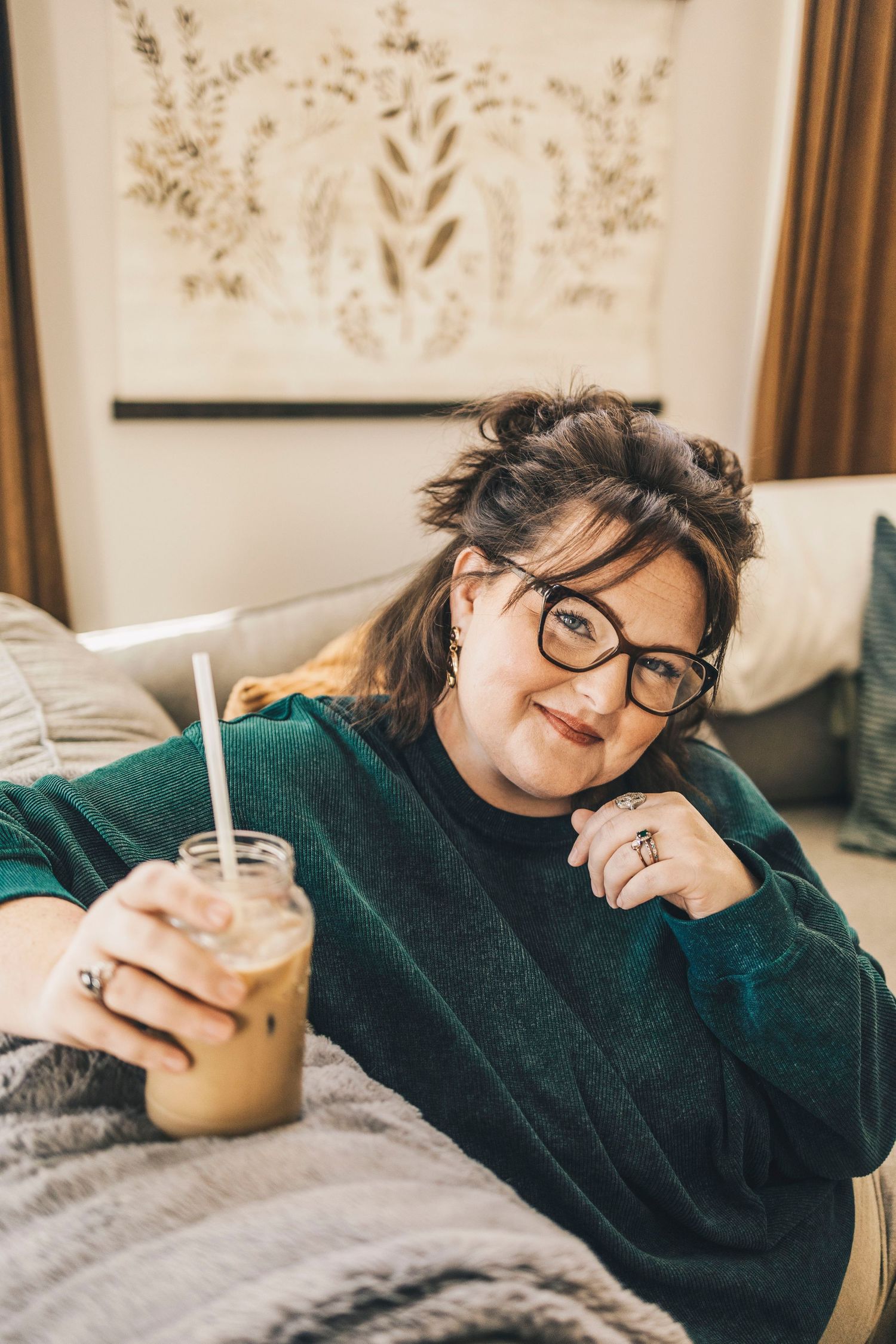 Woman with glasses in green sweater relaxing on bed with iced coffee.