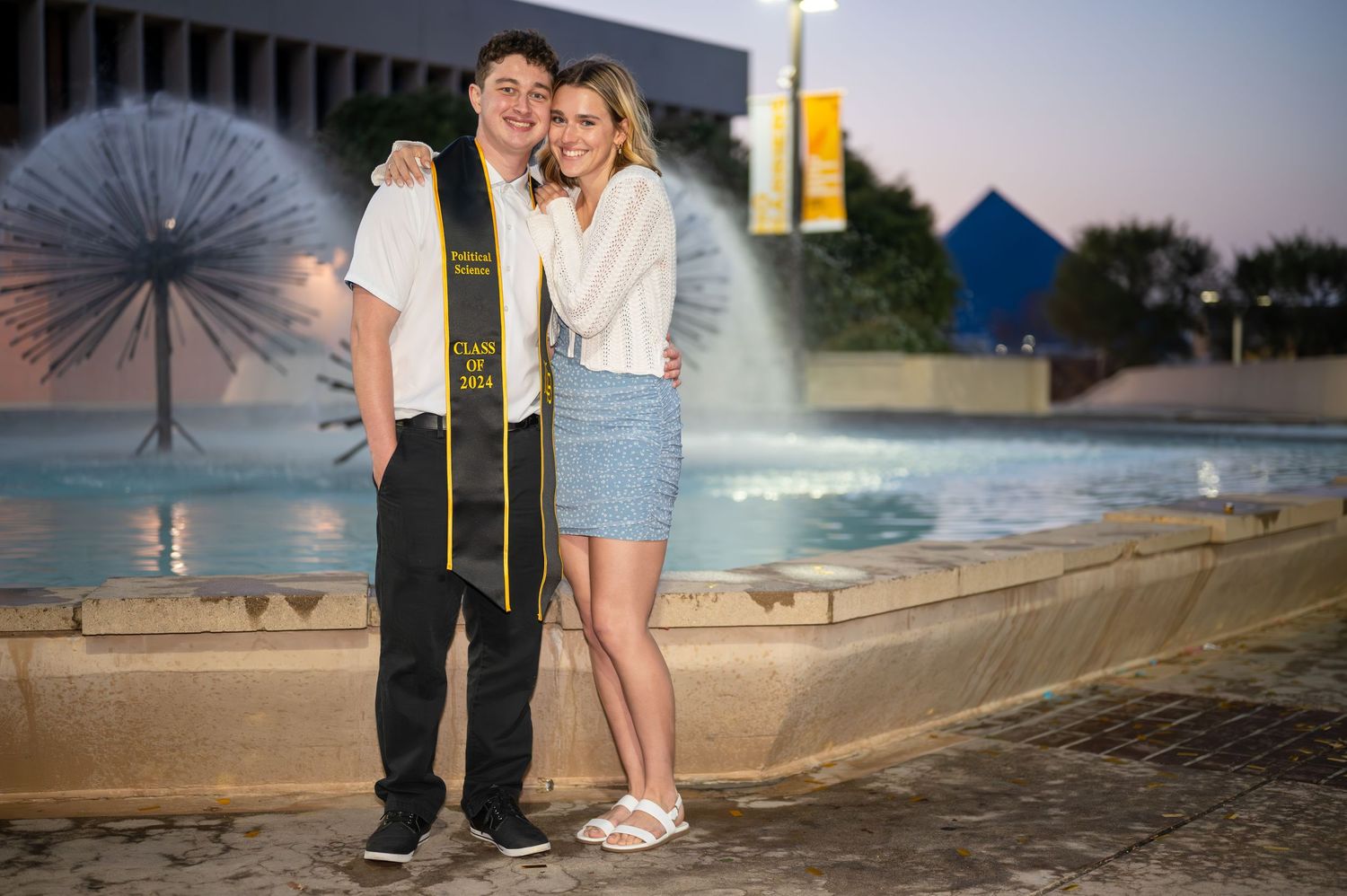 A couple stands together near a water fountain at dusk with lighting equipment visible in the background.